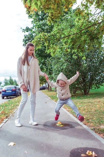 Жінка мама гуляє зі своєю дитиною, восени в місті, маленький хлопчик стрибає через калюжі на тротуарі. Відпочиньте на прогулянку на вихідні. Повсякденний теплий одяг . — стокове фото