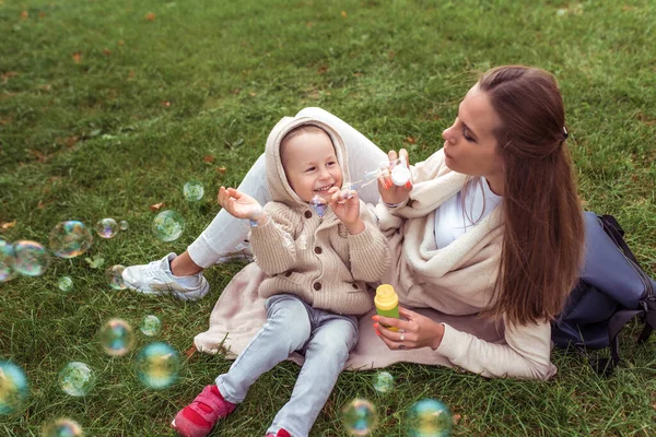 Šťastný malý chlapec hraje a praskne mýdlové bubliny, žena maminka nafoukne mýdlové bubliny. Podzimní den v létě na trávě. Víkendové přestávky, péče o rodiče a péči o děti. Zábavný smích a radost. — Stock fotografie