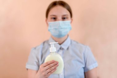 12-15 yaşlarında, tıbbi maske takmış bir kız elinde antiseptik, sıvı sabun, bakım ve hastalara güvenliği ve ellerini yıkamayı hatırlatır..