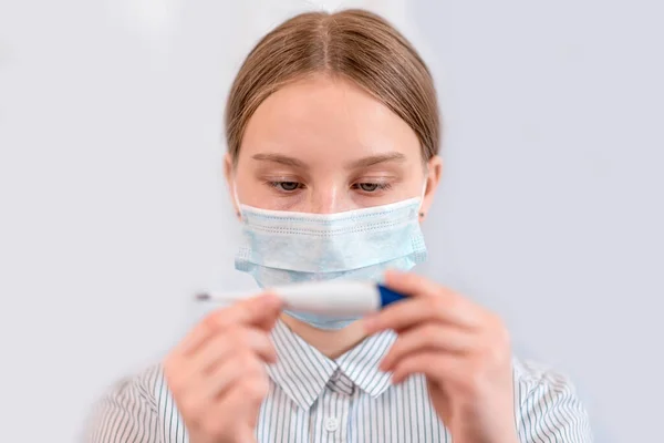 Gadis remaja, topeng medis menutupi wajahnya, memegang tangan termometer elektronik digital, suhu pemeriksaan, gejala penyakit, komplikasi flu, wabah virus pandemi konsep perlindungan virus. Stok Gambar