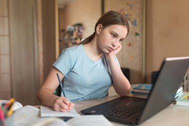 Yorgun bir kız 12-15 yaşlarında, evde kalemle yazıyor, karatahta dersleri yazıyor, internette öğreniyor, bilgisayarında internet var. Karantina süresince uzak eğitim, evde kalın.