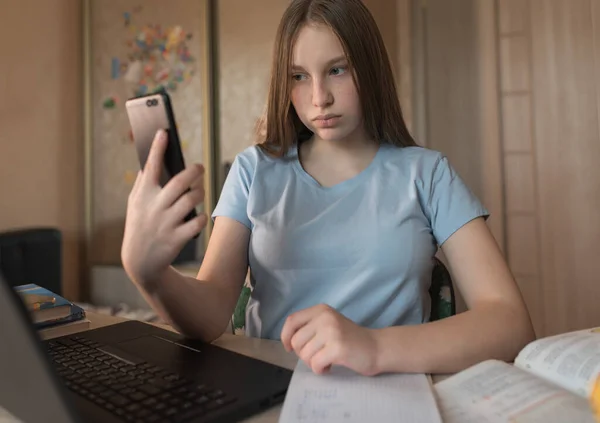 Menina adolescente grava chamada de vídeo no telefone, em casa faz lição de casa na Internet, no laptop, em um smartphone 4G 5G conexões sociais. Ensino à distância e formação em quarentena, ficar em casa . — Fotografia de Stock