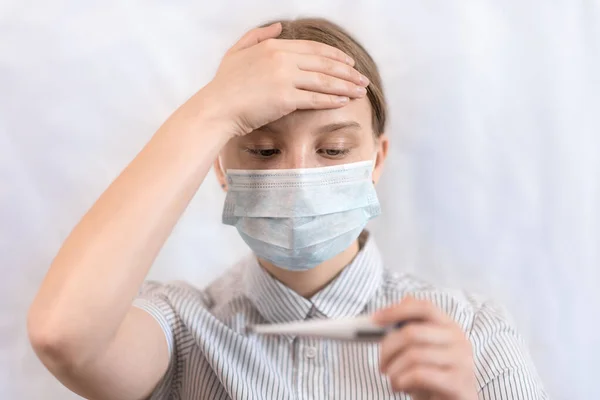 Κοντινό πλάνο πορτρέτο, έφηβο κορίτσι με προστατευτική ιατρική μάσκα, αγγίζει το μέτωπό της με το χέρι της, κρατά έλεγχο θερμοκρασίας στο ηλεκτρονικό θερμόμετρο στο χέρι της. Μείνε σπίτι, ιός και πανδημία. — Φωτογραφία Αρχείου