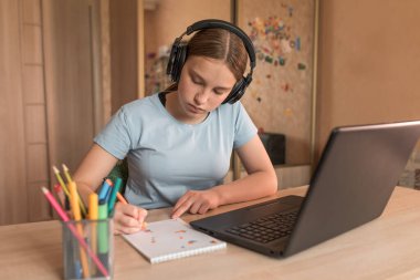 12-14 yaşlarında genç bir kız. Laptop kulaklığı, ödevi, not defteri boyalı kalemleri var. E-eğitim mesafesi internet video iletişimi öğreniyor. Sınav sınavının başarısı.