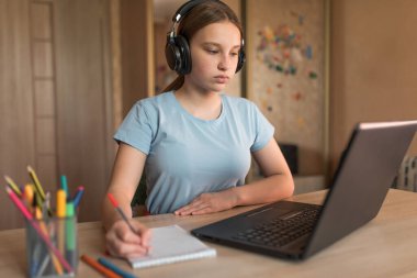 12-14 yaşlarında ciddi bir genç kız, ev kulaklığı, dizüstü bilgisayar dersleri, not defteri boyalı kalemler. E-eğitim mesafesi internet video iletişimi öğreniyor. Sınav sınavının başarısı.