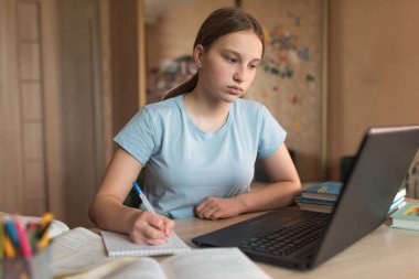 12-14 yaşlarındaki konsantre genç kız, ev, dizüstü bilgisayar dersleri, defter kalem yazıyor. E-eğitim mesafesi internet ve video iletişimi öğreniyor. Ders başarısı ve sınav sınavı.