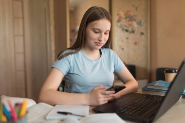 Mutlu gülümseyen kız, evde akıllı telefon, dizüstü bilgisayar dersleri. E-eğitim mesafesi internet video iletişimi öğreniyor. İnternet 'te bir mesaj, sosyal ağlar yazar.