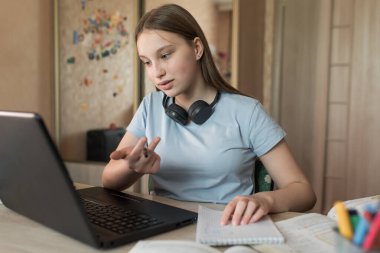 12-15 yaşlarında genç bir kız, öğretmeniyle video bağlantısı kuruyor, ev bilgisayarı derslerinde. E-eğitim ve uzak mesafeden internet video iletişimi öğrenmek. Not defterlerinin kulaklıklarını soyutlar.