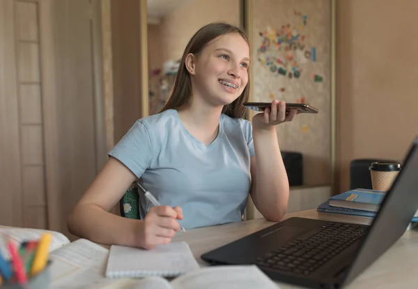 Счастливая улыбающаяся девочка-подросток разговаривает и записывает голосовое сообщение, смартфон, уроки на ноутбуке дома в руке. Электронное образование и дистанционное обучение через Интернет и видеокоммуникации . — стоковое фото
