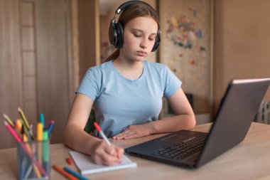 Genç kız not defterinde ders ödevleri yazıyor, evde elektronik eğitim, dizüstü bilgisayar kulaklıkları, internetteki uzaktan video konferansı. Okul eğitimi hazırlık koleji, diploma kursları.