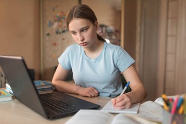 Genç kız not defterine ders ödevleri yazıyor, evde elektronik eğitim, dizüstü bilgisayar kalemi, internetteki uzaktan video konferansı. Okul eğitimi hazırlık koleji, diploma kursları.