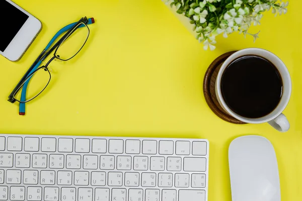 Bright yellow office desk table with keyboard, mouse, a cup of black coffee and eyeglass. Top view with copy space.Working desk table concept.