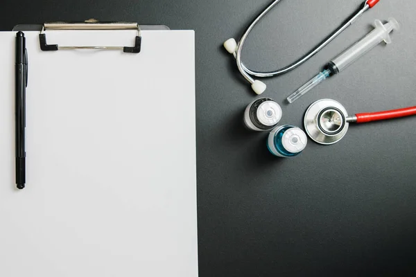 Doctor\'s Day and Covid-19 concept. Clip board with blue and white medical vial, injection and stethoscope on dark table