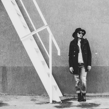 Parlak dudaklı, şık giyinmiş, siyah şapkalı, gözlüklü, siyah ceketli, kot pantolonlu, çelik bir merdivenin yanındaki duvara yaslanmış güzel bir kız.