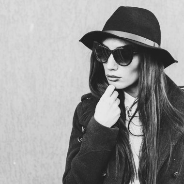 Parlak dudaklı, şık giyinmiş, siyah şapkalı, gözlüklü, siyah ceketli, kot pantolonlu, çelik bir merdivenin yanındaki duvara yaslanmış güzel bir kız.