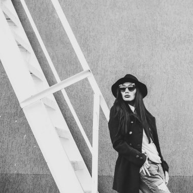 Parlak dudaklı, şık giyinmiş, siyah şapkalı, gözlüklü, siyah ceketli, kot pantolonlu, çelik bir merdivenin yanındaki duvara yaslanmış güzel bir kız.