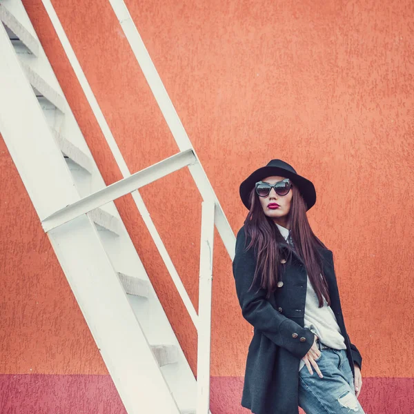 Parlak dudaklı, şık giyinmiş, siyah şapkalı, gözlüklü, siyah ceketli, kot pantolonlu, çelik bir merdivenin yanındaki duvara yaslanmış güzel bir kız.