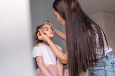 Genç anne kızına makyaj yapıyor, sabah, pencereden gelen ışık, neşeli kız çok konuşuyor.