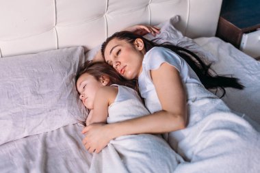 Genç bir anne ve kız sabah aydınlık bir odada uyandılar, yatakta uzanıp birbirlerine baktılar, anne kızını okşadı.