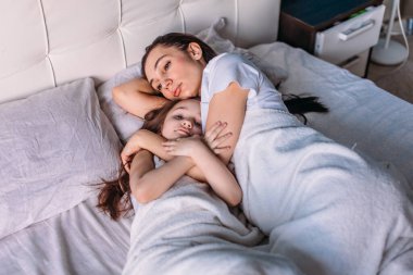 Genç bir anne ve kız sabah aydınlık bir odada uyandılar, yatakta uzanıp birbirlerine baktılar, anne kızını okşadı.