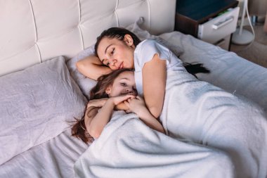 Genç bir anne ve kız sabah aydınlık bir odada uyandılar, yatakta uzanıp birbirlerine baktılar, anne kızını okşadı.