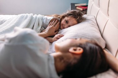 Genç bir anne ve kız sabah aydınlık bir odada uyandılar, yatakta uzanıp birbirlerine baktılar, anne kızını okşadı.