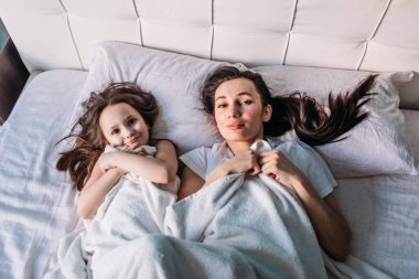 Genç bir anne ve kız sabah aydınlık bir odada uyandılar, yatakta uzanıp birbirlerine baktılar, anne kızını okşadı.