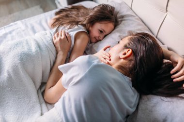 Genç bir anne ve kız sabah aydınlık bir odada uyandılar, yatakta uzanıp birbirlerine baktılar, anne kızını okşadı.