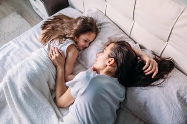 Genç bir anne ve kız sabah aydınlık bir odada uyandılar, yatakta uzanıp birbirlerine baktılar, anne kızını okşadı.