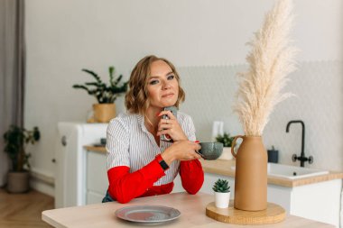 Genç ve güzel bir kız, parlak bir mutfakta, masada oturuyor, fincanına çay ve telefonla mutlu bir şekilde gülümsüyor.