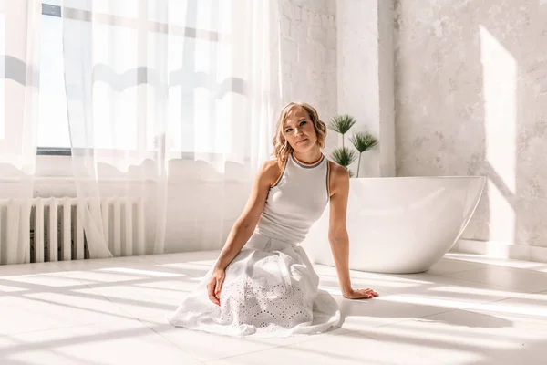 young beautiful girl in a white skirt and white t-shirt, in a spacious bright bathroom, they are in the rays of the sun from a large window, behind it is a white bathtub, she is happy, has a rest, laughs, smiles