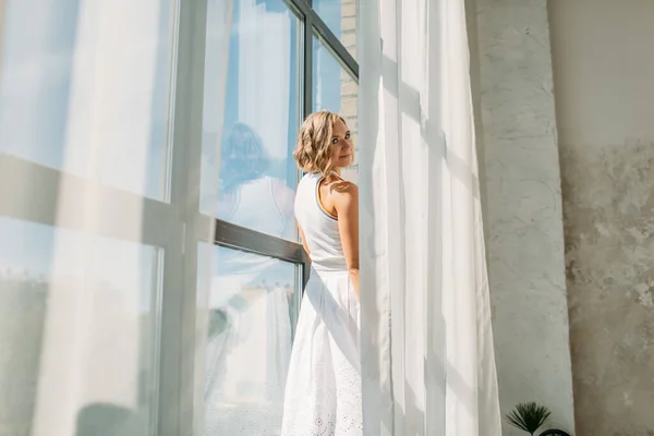 年轻美丽的女孩穿着白色的裙子和白色的T恤 在宽敞明亮的浴室里 站在窗台上 靠近高高的窗户 灿烂的阳光照耀着她 她很快乐 — 图库照片