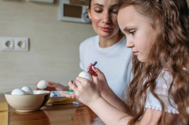 Anne ve kızı sabah mutfakta mutfak masasının üzerinde beyaz yumurtalarla bir fincan oturuyorlar, anne ve kızı beyaz yumurtaların üzerine resim çiziyorlar.