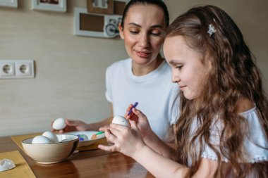 Anne ve kızı sabah mutfakta mutfak masasının üzerinde beyaz yumurtalarla bir fincan oturuyorlar, anne ve kızı beyaz yumurtaların üzerine resim çiziyorlar.