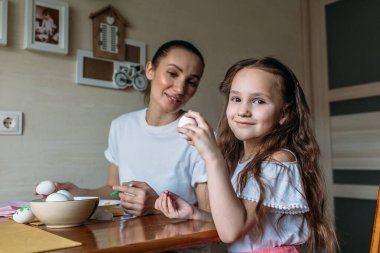 Anne ve kızı sabah mutfakta mutfak masasının üzerinde beyaz yumurtalarla bir fincan oturuyorlar, anne ve kızı beyaz yumurtaların üzerine resim çiziyorlar.