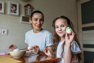 Anne ve kızı sabah mutfakta mutfak masasının üzerinde beyaz yumurtalarla bir fincan oturuyorlar, anne ve kızı beyaz yumurtaların üzerine resim çiziyorlar.