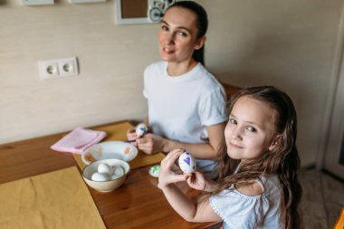 Anne ve kızı sabah mutfakta mutfak masasının üzerinde beyaz yumurtalarla bir fincan oturuyorlar, anne ve kızı beyaz yumurtaların üzerine resim çiziyorlar.