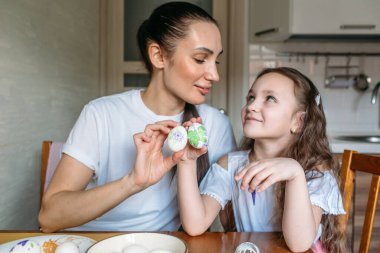 Anne ve kızı sabah mutfakta mutfak masasının üzerinde beyaz yumurtalarla bir fincan oturuyorlar, anne ve kızı beyaz yumurtaların üzerine resim çiziyorlar.