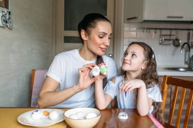 Anne ve kızı sabah mutfakta mutfak masasının üzerinde beyaz yumurtalarla bir fincan oturuyorlar, anne ve kızı beyaz yumurtaların üzerine resim çiziyorlar.