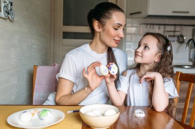 Anne ve kızı sabah mutfakta mutfak masasının üzerinde beyaz yumurtalarla bir fincan oturuyorlar, anne ve kızı beyaz yumurtaların üzerine resim çiziyorlar.