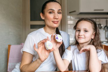 Anne ve kızı sabah mutfakta mutfak masasının üzerinde beyaz yumurtalarla bir fincan oturuyorlar, anne ve kızı beyaz yumurtaların üzerine resim çiziyorlar.
