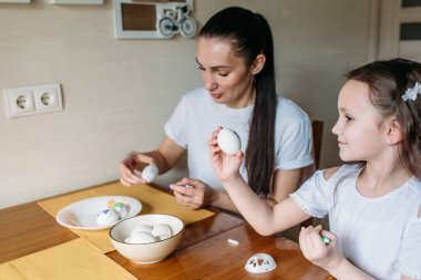 Anne ve kızı sabah mutfakta mutfak masasının üzerinde beyaz yumurtalarla bir fincan oturuyorlar, anne ve kızı beyaz yumurtaların üzerine resim çiziyorlar.