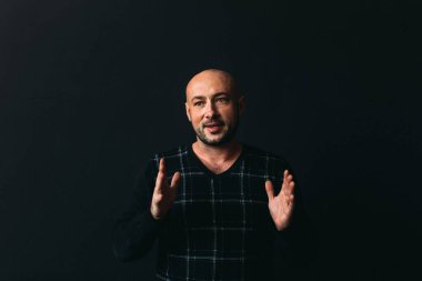 Gözlüklü, koyu renk elbiseli, kel, siyah arka planda genç bir adam elleri ve yüzünde duygular olan bir şey gösterir.