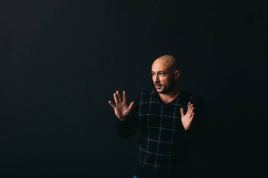 Gözlüklü, koyu renk elbiseli, kel, siyah arka planda genç bir adam elleri ve yüzünde duygular olan bir şey gösterir.