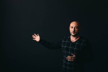 Gözlüklü, koyu renk elbiseli, kel, siyah arka planda genç bir adam elleri ve yüzünde duygular olan bir şey gösterir.