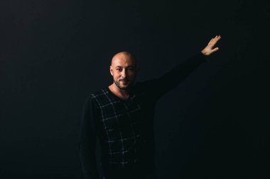 Gözlüklü, koyu renk elbiseli, kel, siyah arka planda genç bir adam elleri ve yüzünde duygular olan bir şey gösterir.