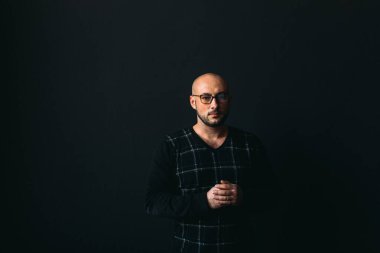 Gözlüklü, koyu renk elbiseli, kel, siyah arka planda genç bir adam elleri ve yüzünde duygular olan bir şey gösterir.
