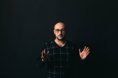 Gözlüklü, koyu renk elbiseli, kel, siyah arka planda genç bir adam elleri ve yüzünde duygular olan bir şey gösterir.