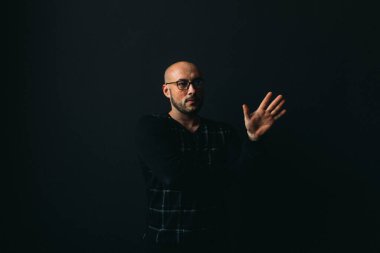 Gözlüklü, koyu renk elbiseli, kel, siyah arka planda genç bir adam elleri ve yüzünde duygular olan bir şey gösterir.