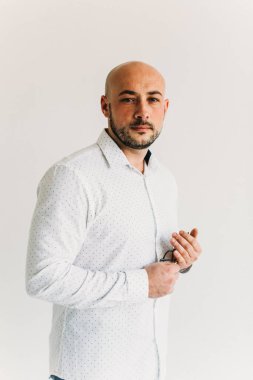 Gözlüklü genç bir adam, beyaz gömlekli, züppe, kel, beyaz arka planda duruyor, düşünceli bir şekilde elinde gözlük tutuyor.